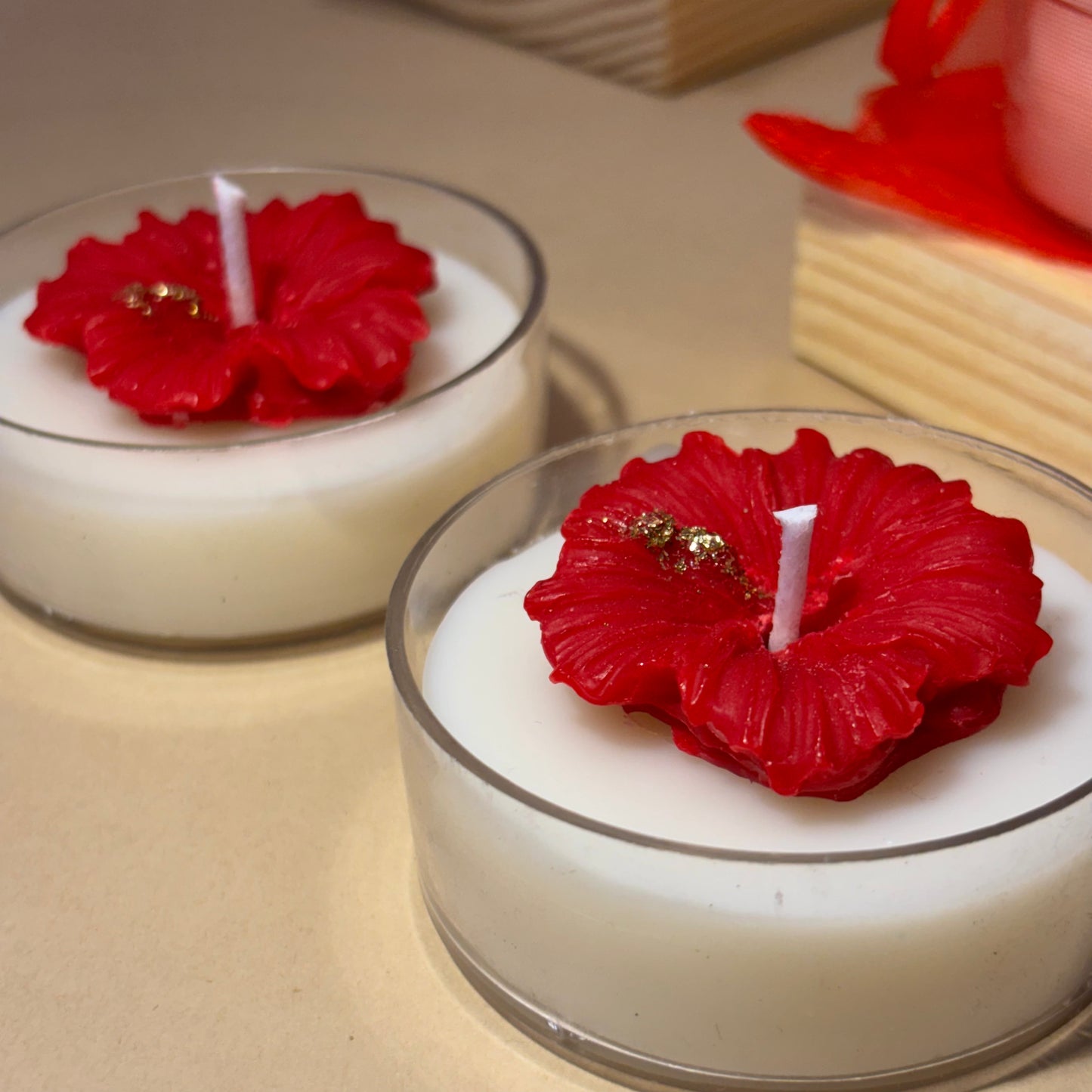 Two white wax candles in clear glass holders, each topped with a red hibiscus flower decoration and a small gold accent, on a beige tabletop.