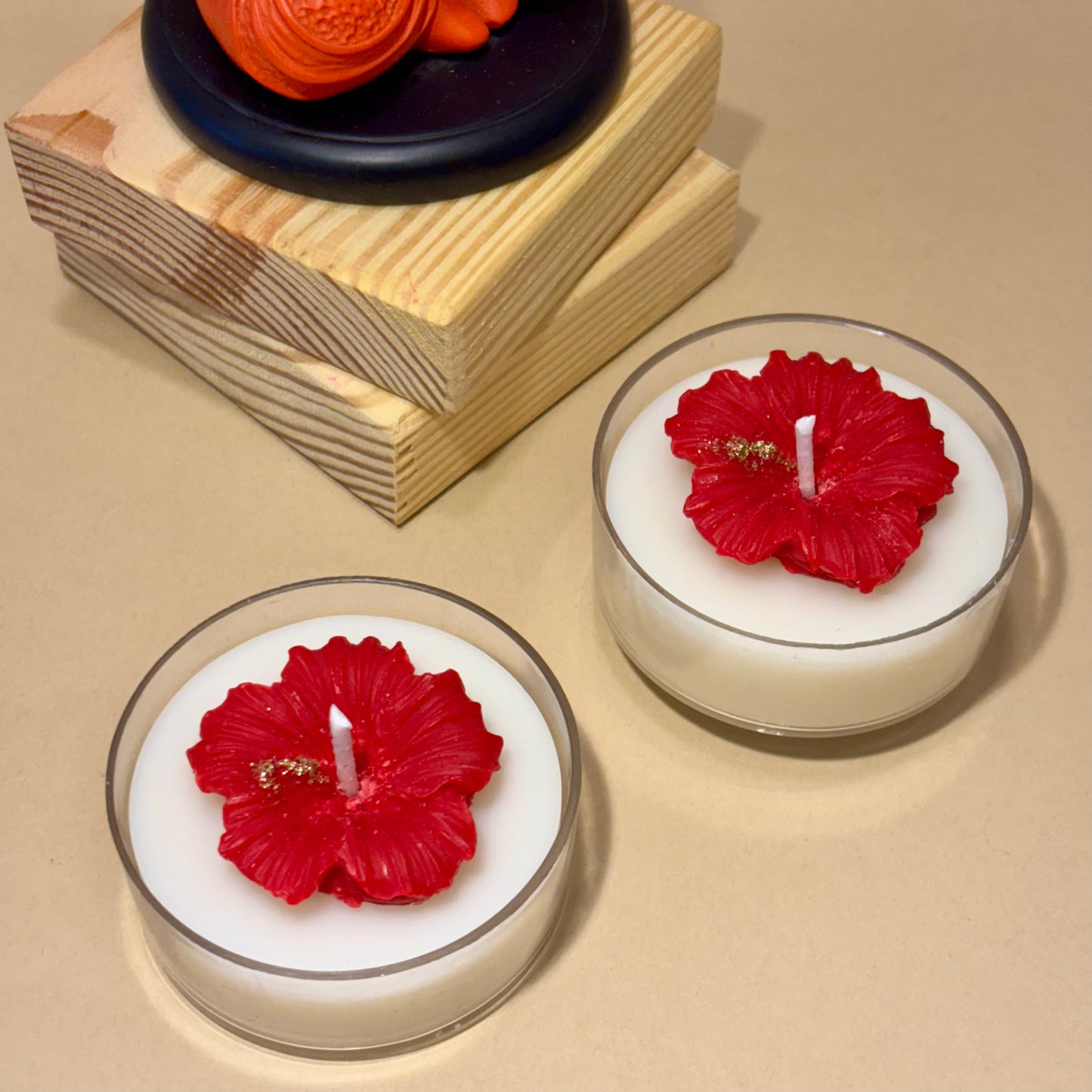 Two white tea light candles with red hibiscus flower tops, displayed on a beige surface with decorative stacked wooden blocks and an orange flower in the background.