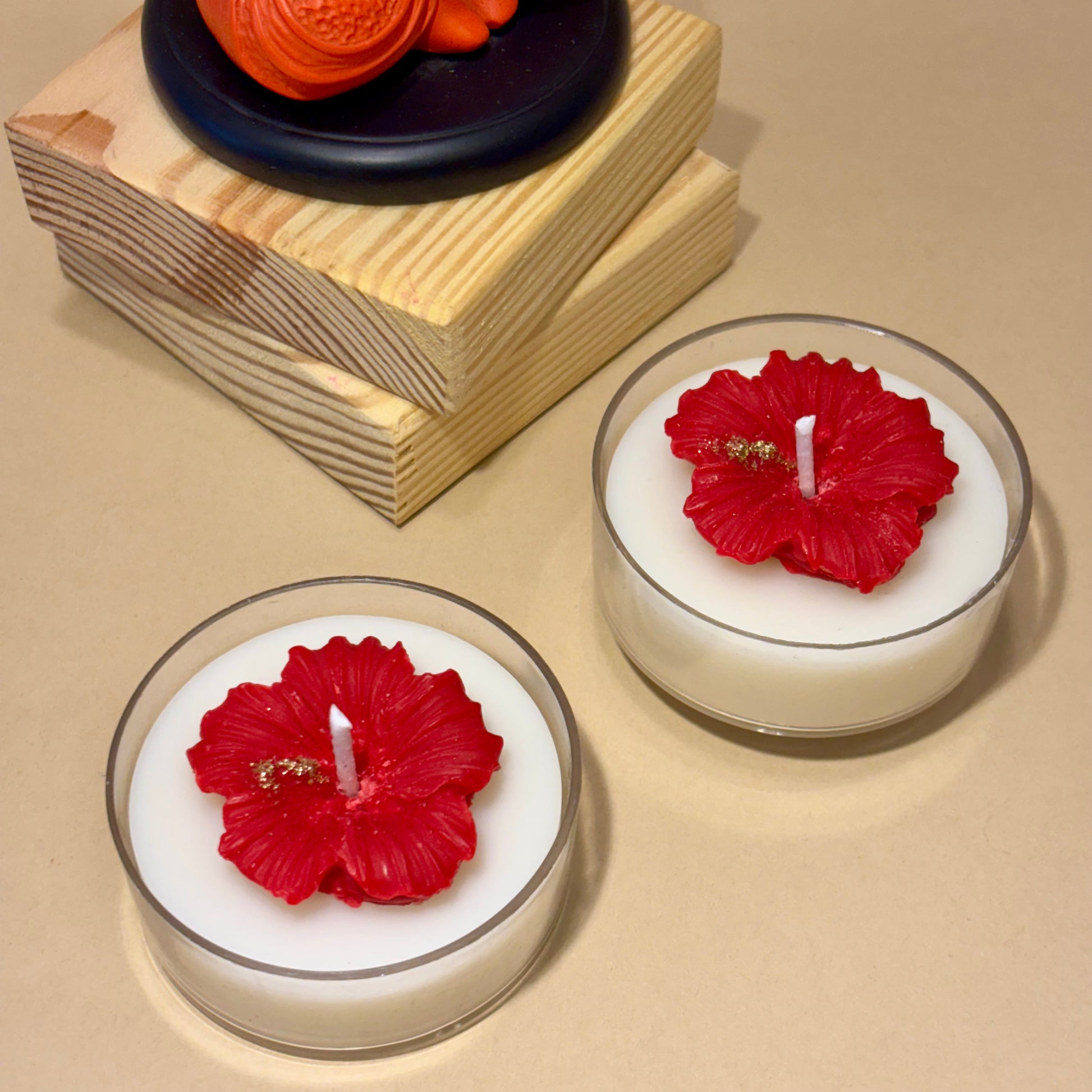Two white tea light candles with red hibiscus flower tops, displayed on a beige surface with decorative stacked wooden blocks and an orange flower in the background.
