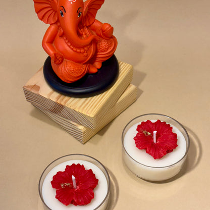 A handcrafted Jaswandi hibiscus candle featuring a red wax flower with gold accents on a white base, styled on wooden blocks next to a vibrant Lord Ganesha idol by Anant The Limitless Art.