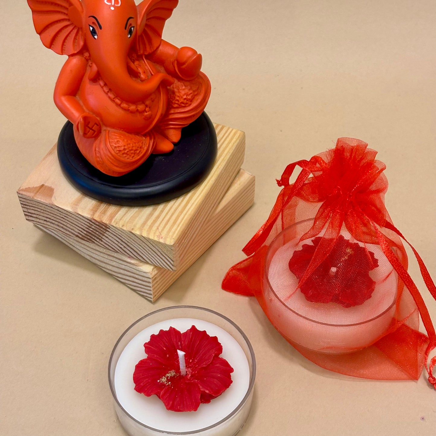 A handcrafted Jaswandi hibiscus candle featuring a red wax flower with gold accents on a white base, styled on wooden blocks next to a vibrant Lord Ganesha idol by Anant The Limitless Art.