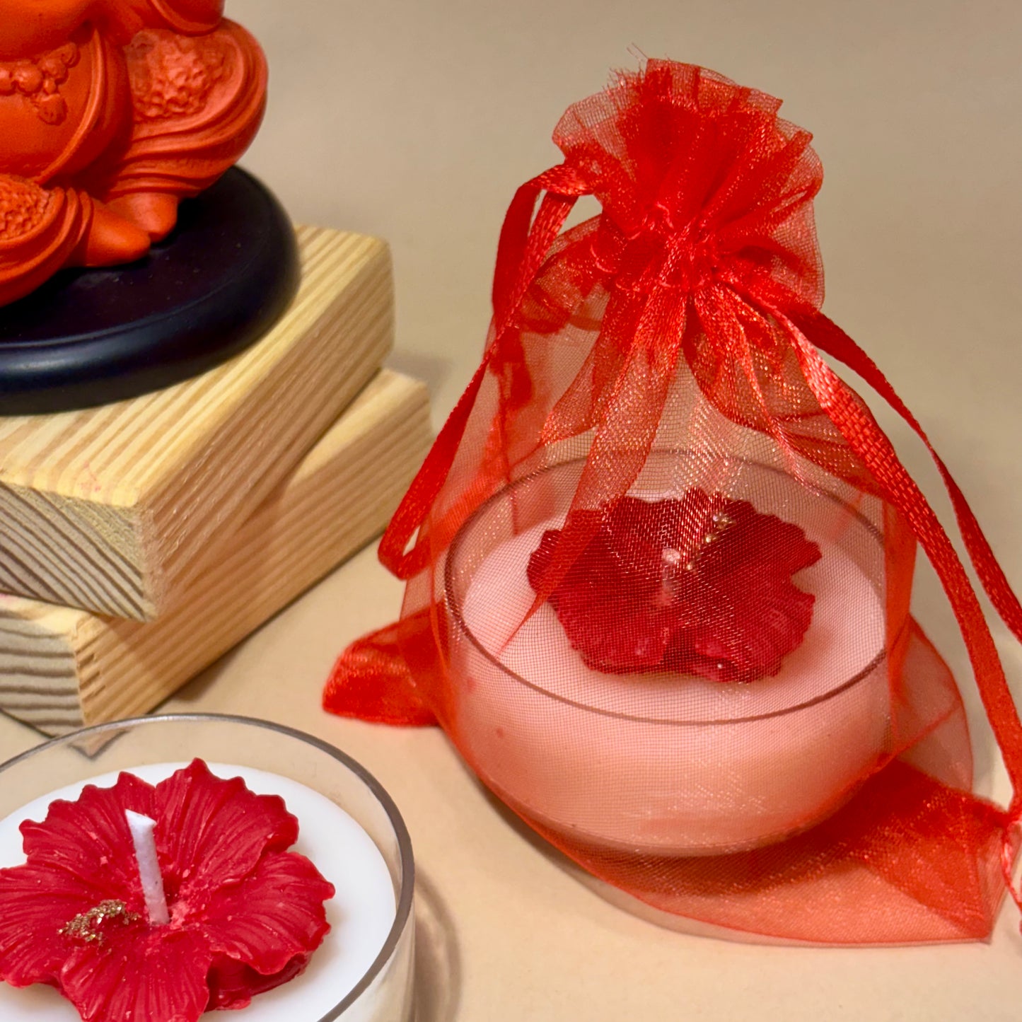 A small pink candle in a clear glass cup tied with a red sheer organza bag and red ribbon, shown beside another red flower-shaped candle on a beige surface with decorative blocks in the background.