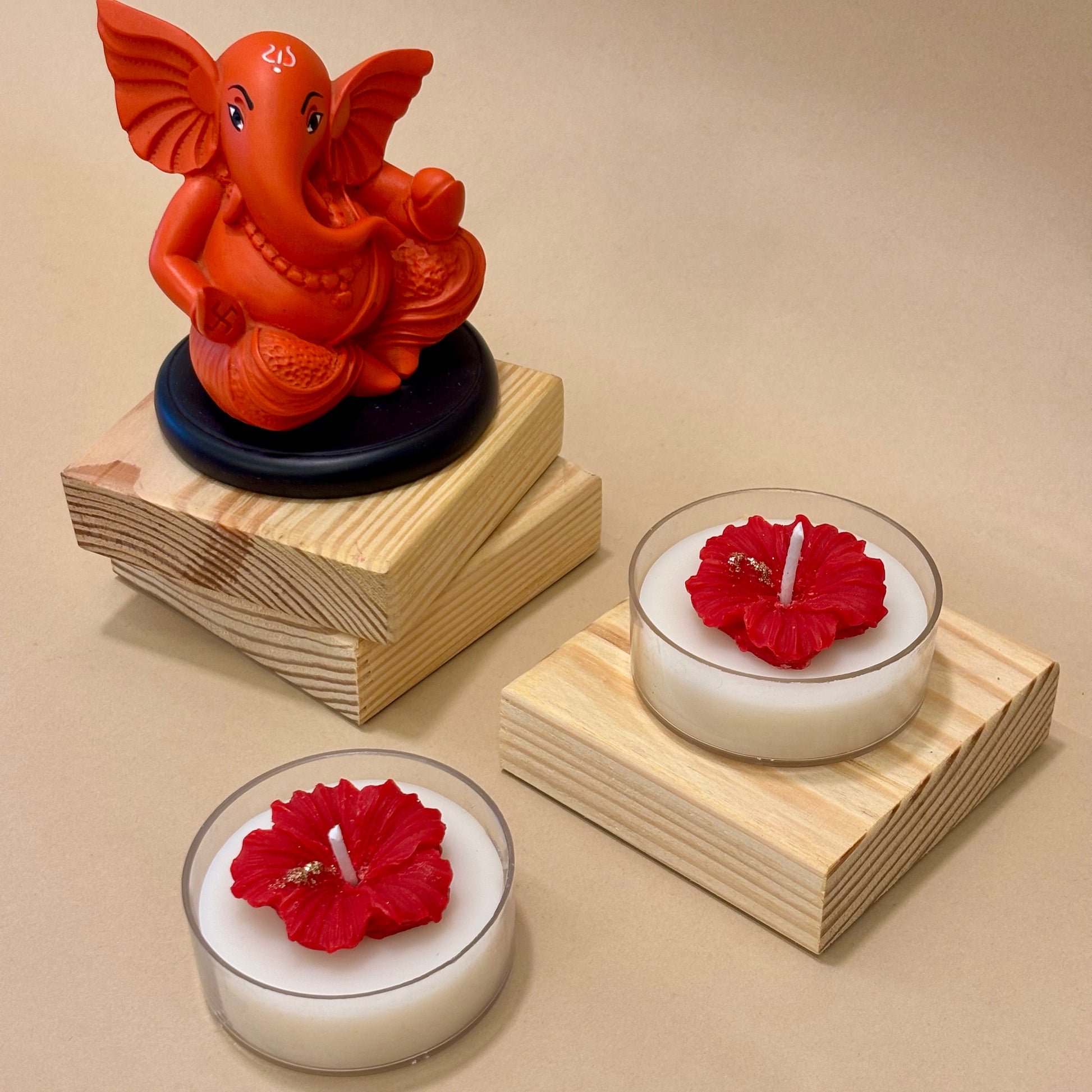 A handcrafted Jaswandi hibiscus candle featuring a red wax flower with gold accents on a white base, styled on wooden blocks next to a vibrant Lord Ganesha idol by Anant The Limitless Art.