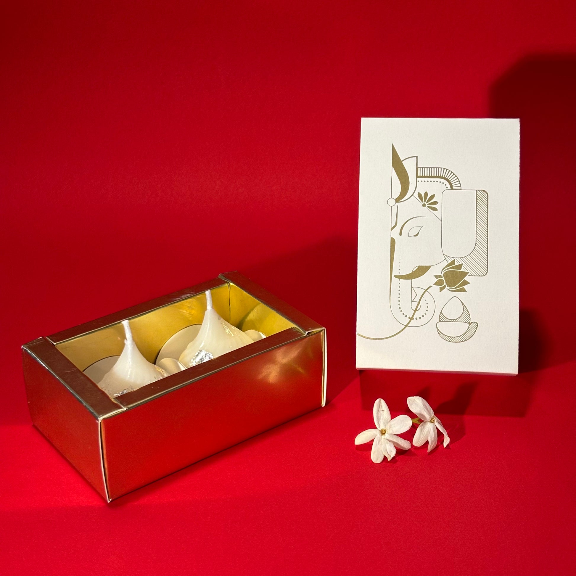 A boxed set of Modak candles in a metallic gold box, shown beside a white decorative card with gold line art and two small white flowers on a bright red background.