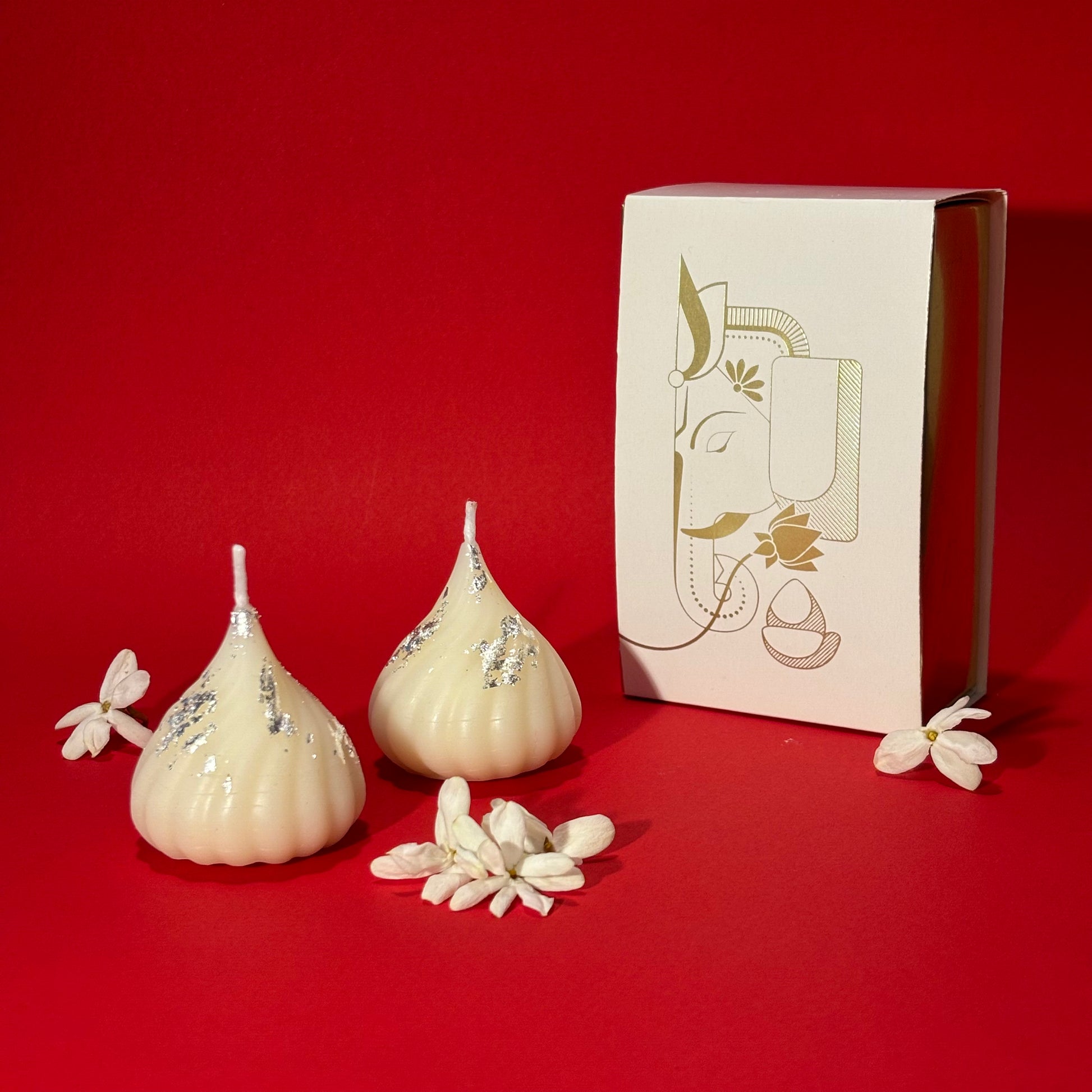 Two cream-colored Ganesha-shaped candles with silver foil accents are displayed next to their white packaging box featuring a gold-toned Ganesha illustration, set against a solid red background.