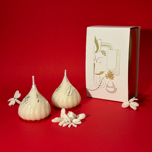 Two cream-colored Ganesha-shaped candles with silver foil accents are displayed next to their white packaging box featuring a gold-toned Ganesha illustration, set against a solid red background.